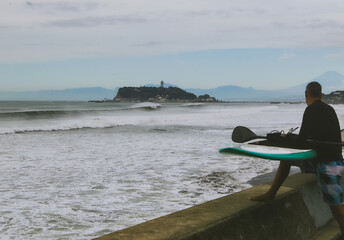 Stand Up Paddle Boarding In Japan near to Enoshima Island with My Fuji in the background. A foreign man with his board looking out to sea where large waves are breaking.