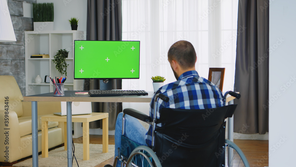 Poster invalid man in wheelchair working on computer with green screen.