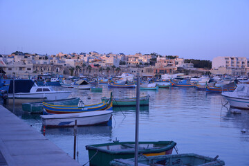 Fototapeta na wymiar Multi-colored fishing boats rest in the sea bay against the background of a small old golden city and a blue cloudless sky.