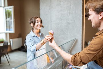 Young and cheerful woman buying ice cream in waffle cone, standing with a seller at the counter of a shop