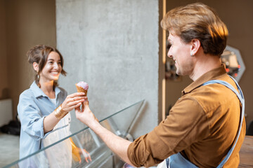 Young and cheerful woman buying ice cream in waffle cone, standing with a seller at the counter of...