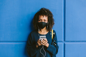 Portrait of urban afro hairstyle woman wearing face mask and using her phone for listening music. Coronavirus Covid-19 new lifestyle.