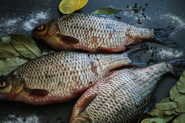 Three river crucians prepared for frying on a wooden table with seasonings and lemon slices.
