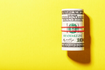 A roll of hundred-dollar American bills is tied with a red elastic band on a yellow background.