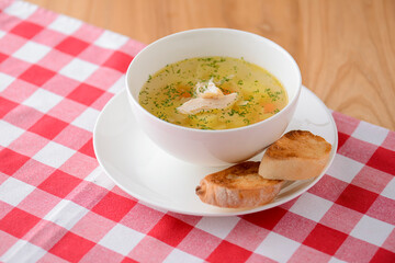 Chicken soup with vegetables and meat served in a white bowl over rustic wooden background with red plaid tablecloth.