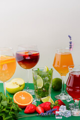 Five tropical mixed drinks, orange, lemon and raspberries cocktails over bright pastel green background.
