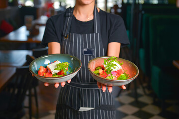 Close up of waiter serving two plates of vegetable salad with feta cheese, lettuce and tomatoes. Restaurant service.