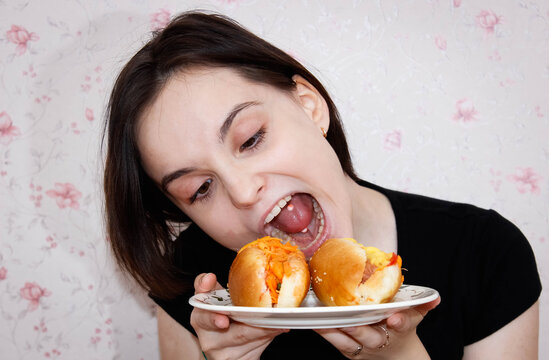 A girl with a piercing in her tongue and a grimace on her face tries to bite off a hot dog on a plate. - Powered by Adobe