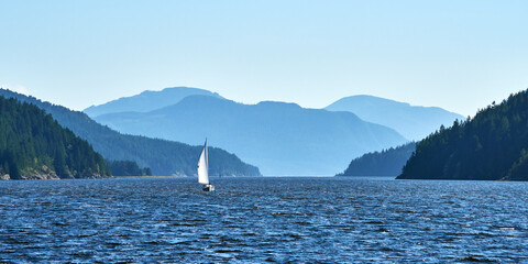 A panoramic view of the mountains around the ocean bay and a lone sailboat near Bamfield in Canada.