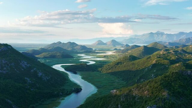 River meandering through the valley toward distant mountains, in the morning. Crnojevica river in Montenegro, flowing through the marsh, among hills, on its way to lake Skadar. Aerial footage.