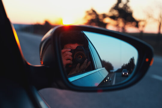 Reflection in car mirror of young guy taking photo of sunset with digital camera.
