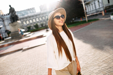 Fototapeta premium Carefree beautiful long haired girl in hat and sunglasses is laughing and looks away, she is walking in downtown. Summer portrait in warm natural sunlight