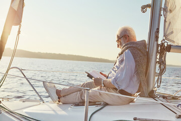 Relaxed senior man reading a book, while sitting on the side of sail boat or yacht deck floating in...