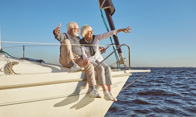 A happy senior couple sitting on the side of a sail boat on a calm blue sea, pointing at landscape, enjoying view
