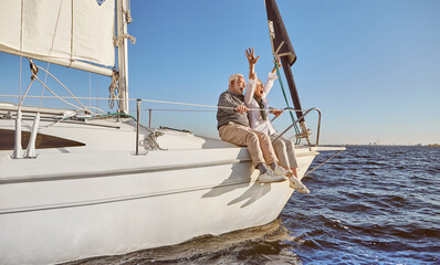 A happy senior couple sitting on the side of a sail boat on a calm blue sea, enjoying view. Woman...