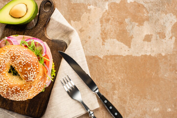 top view of fresh delicious bagel on wooden cutting board on aged beige surface with avocado, cutlery and napkin