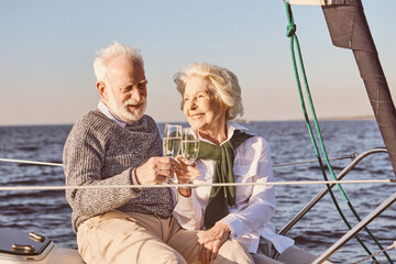 Fototapeta na wymiar Cheers. Close up of happy senior couple sitting on the side of sail boat or yacht deck floating in sea. Man and woman drinking wine or champagne, enjoying the view