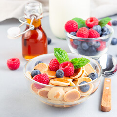 Tiny cereal pancakes served with maple syrup, raspberry, blueberry and milk, in a glass bowl, square