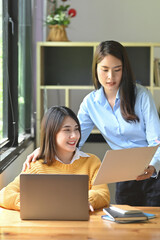 Two women are working together with a computer laptop at the wooden working desk.