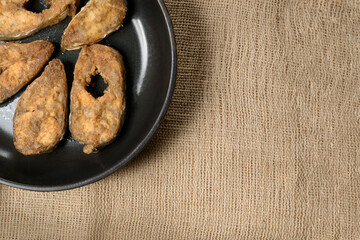 Slices of fried fish fillets in a pan, burlap background. View from above.