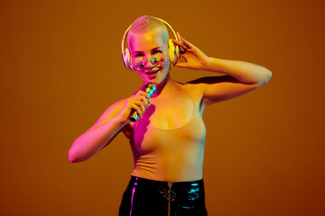 Musician. Young caucasian woman on brown studio background in neon light. Beautiful female model with microphone, speaker. Human emotions, facial expression, sales, ad concept. Freak's culture.