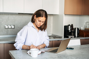 Concentrated young woman freelancer is working with a laptop from home at a table in the kitchen. Remote Work Concept