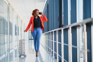 Young african american female passanger in casual clothes is in airport with baggage. Using phone. Travel concept