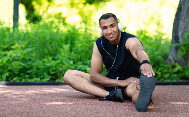 Portrait of young black man in headphones stretching his legs on jogging track at park, copy space