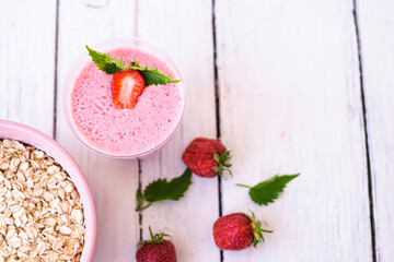 Cocktail with strawberries and milk in a glass decorated with mint and strawberries. Nearby is a pink cup with Hercules flakes, and loose strawberries on a white wooden background. 