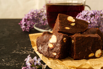 A piece of homemade chocolate cake on crumpled craft paper, with nuts on a black texture table. A cup of tea and lilac flowers decorate a delicious tea party.