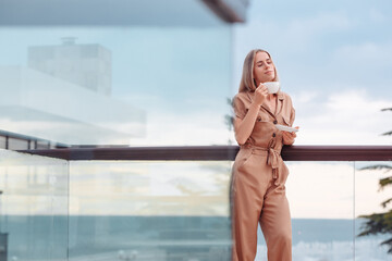 Portrait of a happy smiling caucasian woman in stylish overalls with a cup of coffee meeting the day standing on the hotel terrace with a gorgeous view of the sea and nature