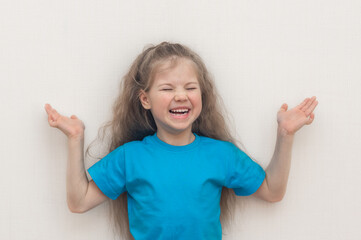 Portrait of a funny blond little girl in a blue T-shirt who smiles and laughs isolated on a white (light) background. Hands up. copy space.