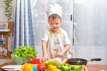 Cute happy smiling Asian little boy child wearing chef hat and apron having fun preparing, cooking healthy food in kitchen, Fun indoor activities for kindergarten kids, photo in real life interior