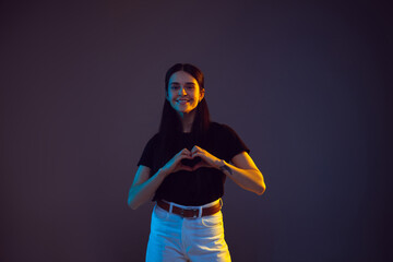 Heart shaped hands. Caucasian young woman's portrait on dark studio background in neon. Beautiful brunette model in casual. Concept of human emotions, facial expression, sales, ad, love. Copyspace.