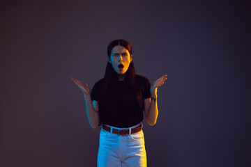 Angry, shocked. Caucasian young woman's portrait on dark studio background in neon. Beautiful female brunette model in casual. Concept of human emotions, facial expression, sales, ad. Copyspace.