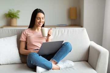 Happy Woman Using Laptop Enjoying Coffee Sitting On Sofa Indoor