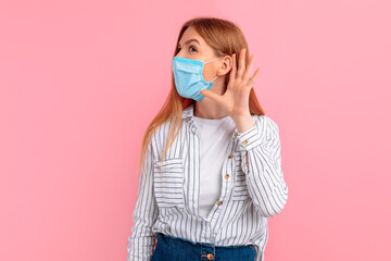 curious, focused woman in a medical protective mask on her face, listening with her hand to her...