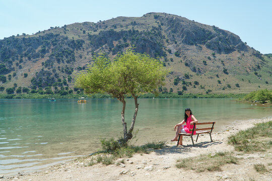 A girl is sitting on a bench near a tree on lake kurnas. Island of Crete.