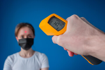 Man hand holding yellow pyrometer and measuring temperature of woman in medical face mask looking...
