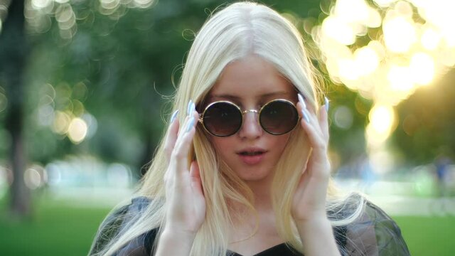 Attractive young blonde girl with long hair and black dress in park gracefully lowers sunglasses, winks playfully and looks with alluring look, then puts glasses back on, close-up view in slow motion.