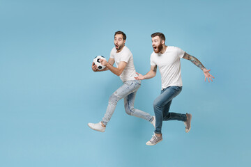 Fototapeta na wymiar Shocked two young men guys friends in white t-shirt isolated on pastel blue background in studio. Sport leisure lifestyle concept. Cheer up support favorite team with soccer ball jumping like running.