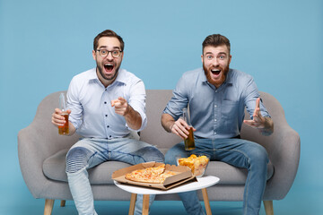 Amazed men guys friends colleagues in casual shirt sit on couch isolated on pastel blue background. Sport leisure concept. Cheer up support favorite team hold beer bottle point index finger on camera.