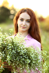 Naklejka na ściany i meble A young beautiful red-haired girl close-up is looking at the camera with a huge bouquet of field daisies in a pink dress. Sunset on a summer evening. Effect bokeh, flash.