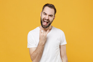 Funny young bearded guy in white casual t-shirt posing isolated on yellow background studio portrait. People lifestyle concept. Mock up copy space. Depicting heavy metal rock sign, horns up gesture.