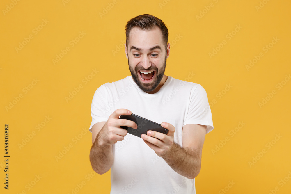 Wall mural Excited young bearded man guy in white casual t-shirt posing isolated on yellow wall background studio portrait. People lifestyle concept. Mock up copy space. Play game with mobile phone, screaming.