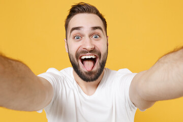 Close up of cheerful young bearded man guy in white casual t-shirt posing isolated on yellow background. People lifestyle concept. Mock up copy space. Doing selfie shot on mobile phone showing tongue.