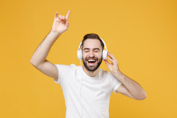 Fototapeta premium Funny young bearded man guy in white casual t-shirt posing isolated on yellow background studio. People lifestyle concept. Mock up copy space. Listen music with headphones keeping eyes closed dancing.