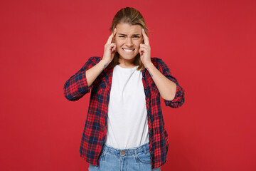 Dissatisfied tired young blonde woman girl in casual checkered shirt posing isolated on red background studio portrait. People lifestyle concept. Mock up copy space. Put hands on head having headache.