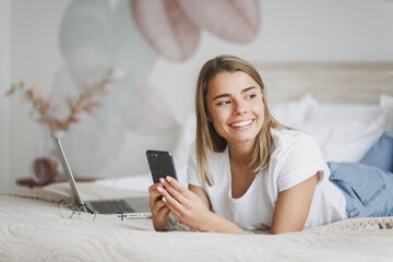 Smiling young woman girl in white t-shirt lying in bed with white sheet pillow blanket spending time in bedroom at home. Rest relax good mood lifestyle concept. Using mobile phone, typing sms message.