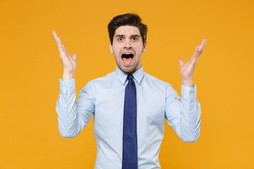 Shocked irritated young business man in classic blue shirt tie posing isolated on yellow background studio. Achievement career wealth business concept. Mock up copy space. Spreading hands, screaming.